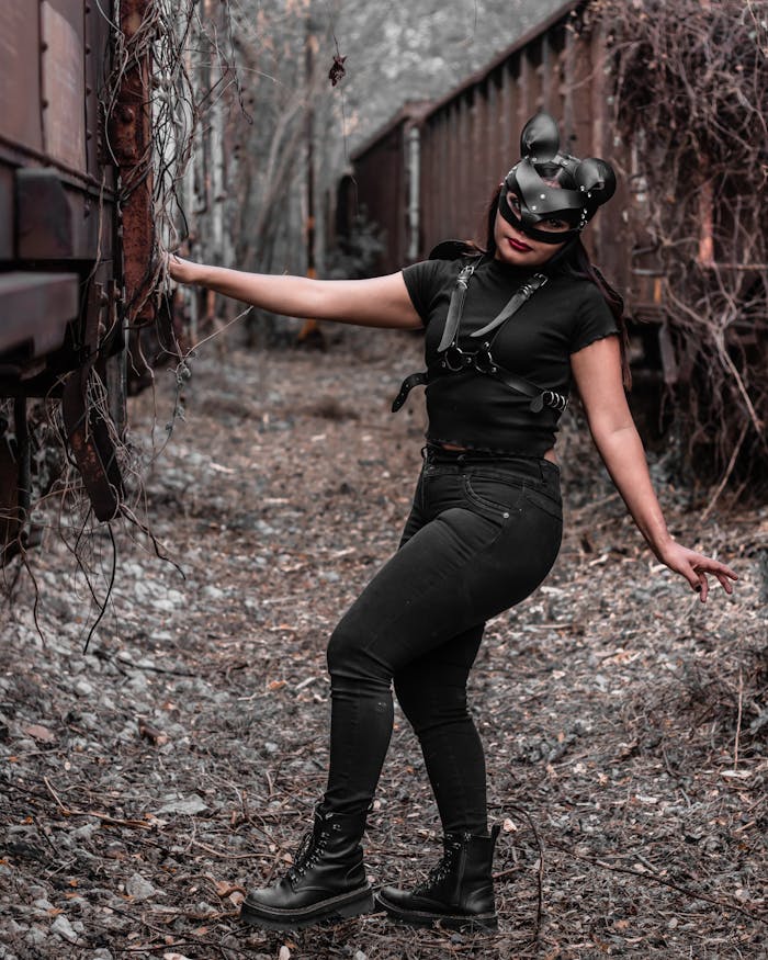 Model in black leather outfit posing with mask beside abandoned cargo trains outdoors.