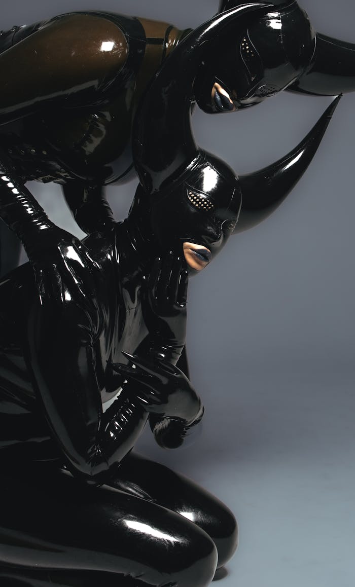 Two women in glossy black latex with horned masks, posing in a studio setting.