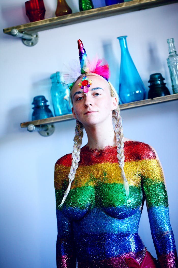 Portrait of a woman in rainbow glitter body paint with braided hair and unicorn headpiece indoors.